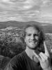 Joshua Zimmerman. A white man with curly hair standing on top of the mountain. He is making a rock sign with his left hand in front of his face.